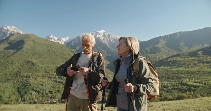 Old couple having a hike in spring mountains, then stopping to take a picture on a smartphone. Senior caucasian family spending time together travelling after retirement - tourism concept 4k