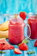 In glasses, freshly squeezed juice from strawberries, watermelon, banana and mint next to a strawberry berry piece of watermelon ljyatyat and banana on a wooden blue background