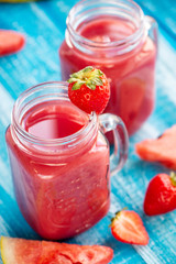In glasses, freshly squeezed juice from strawberries, watermelon, banana and mint next to a strawberry berry piece of watermelon ljyatyat and banana on a wooden blue background