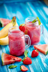 In glasses, freshly squeezed juice from strawberries, watermelon, banana and mint next to a strawberry berry piece of watermelon ljyatyat and banana on a wooden blue background