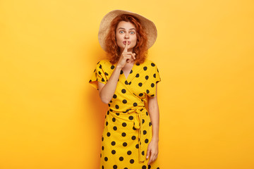 Cute young girl shows shush gesture with surprised expression, keeps index finger over mouth, has ginger wavy hair, dressed in fashionable polka dot dress, tells secret, isolated on yellow studio wall