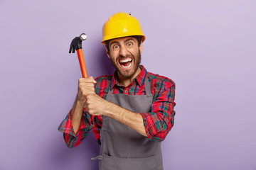 Desperate male laborer or repairman holds hammer with both hands, has outraged facial expression,...