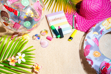 Fototapeta na wymiar Colourful writing note pad with pink and green markers on sand, surrounded by green palm leafs, sea shells, pink hat, inflatable toys. Summer beach background, flat lay