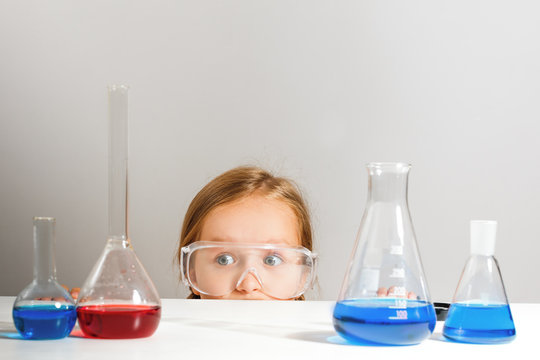 A little girl in safety glasses is hiding in a chemistry lab. The child looks out from under the table.