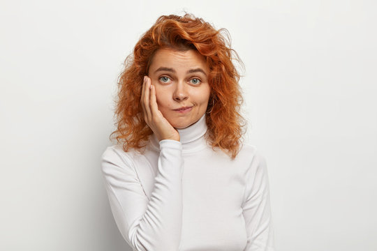 Close up shot of lovely woman with red curly hair, purses lips and looks with doubtful expression, touches cheek, poses against white background, wears poloneck jumper. Human facial expressions