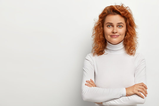 Portrait of good looking redhead woman looks with little smile, has calm face expression, keeps arms folded, wears white turtleneck, poses alone, listens pleasant comments, isolated on white wall