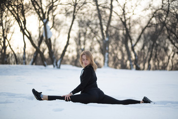 Young caucasian female blonde in leggings stretching exercise sitting on a string at open air in snowy forest. fit and sports lifestyle