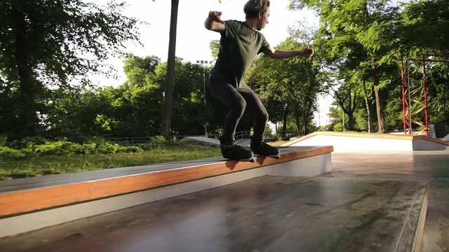 Chasing footage of a young rollers skater sliding on a wooden railings in the mordern skate park with ramps. Sunny, summer day. Slow motion. Close up