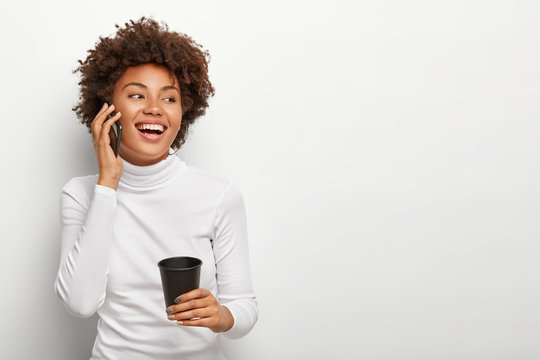 Photo of satisfied carefree woman with curly haircut, talks via smartphone, looks positively aside, drinks takeout coffee, being in good mood during lively conversation. People and lifestyle