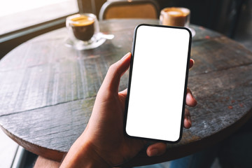 Mockup image of a hand holding and showing black mobile phone with blank white screen in cafe