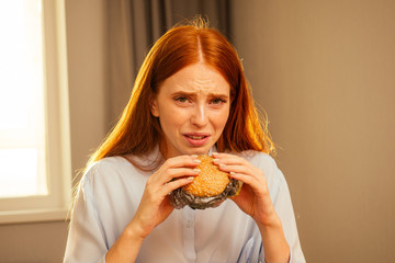 redhaired ginger girl eating fake burger with bun sesame and plastic cellophane film trash,drinking water from disposable cup with non-eco tube at home kitchen