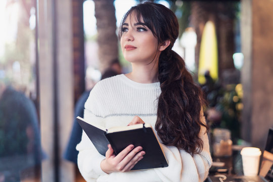 Portrait Of Young Brunette Woman Standing In Cafe, Writing In Notebook, Looking Out Window. Girl Makes Notes In Diary, Plans.