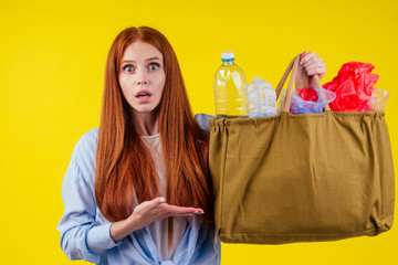 redhead ginger girl with consternation on her face pointing finger copyspase with bad full of non-eco plastic trash in studio yellow background