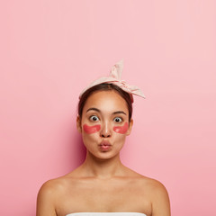 Adorable Asian woman applies self sticking patches under eyes to reduce puffiness and dark circles, wears headband on head, keeps lips folded, stands bare shoulders, isolated on pink background