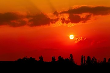 Dark red and orange sunset sky with clouds and black horizon with trees. Summer dusk landscape.