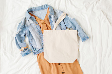 Blue jean jacket and  beige dress with tote bag on white bed. Women's stylish autumn outfit. Trendy clothes with white eco bag mockup. Flat lay, top view.