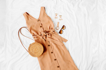 Beige dress with bamboo bag and accessories on white bed. Women's stylish autumn or summer outfit....