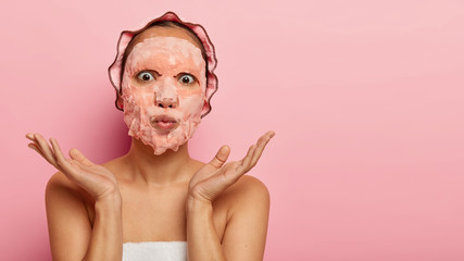 Surprised woman has cosmetic tissue mask on face, stares with eyes popped out, lips folded, spreads palms sideways, enjoys spa procedures after taking shower, wants to be healthy and beautiful