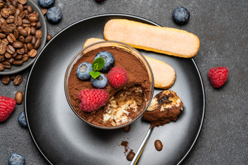 Classic tiramisu dessert portion with blueberries and raspberries in a glass cup on stone serving board on concrete background