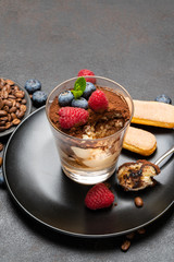 Classic tiramisu dessert portion with blueberries and raspberries in a glass cup on stone serving board on concrete background