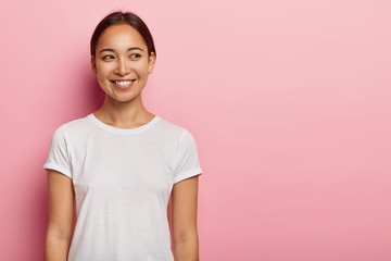 Studio shot of happy young Asian woman has tender smile, looks aside with charming expression,...