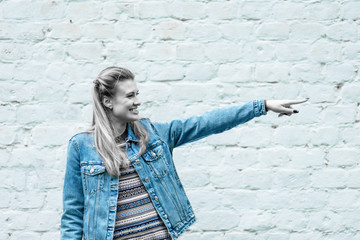 Young attractive woman in casual jeans clothes at a white brick wall points outstretched hand direction