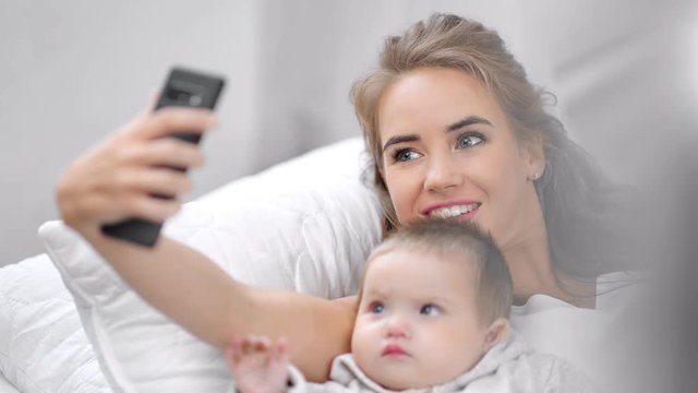 Smiling attractive young mother taking selfie posing with cute baby using smartphone close-up