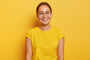 Horizontal shot of happy Asian female student wears big round spectacles, yellow casual wear,...
