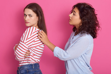 Beautiful girls with dark hair, lady wears striped shirt keep arms folded, do not talk to her friend, woman dressed blue shirt touches female's shoulders, wants to talk, pose isolated over pink wall.