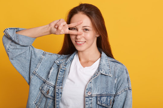 Smiling attractive woman with dark brown hair, showing two fingers forming the letter V like vicory, covering her eye with piece gesture, wearing denim jacket and white t shirt , isolated over yellow.