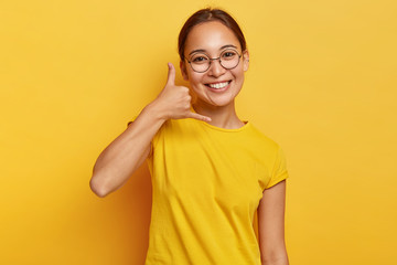 Smiling delighted Asian girl shows call me gesture, makes phone hand sign, has happy expression,...