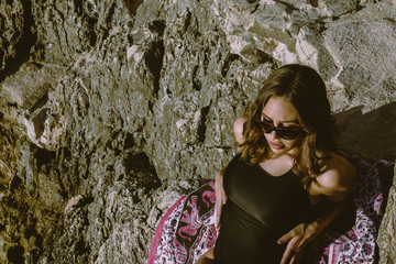 Young hipster woman lying on the rocks in a swimsuit over beach towel. Young woman lying near the sea with vintage sunglasses.