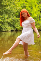 happy redheaded woman in white dress at a river having fun and splashing water in summer. happy person plays with water 