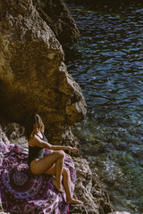 Young hipster woman lying on the rocks in a swimsuit over beach towel. Young woman lying near the sea with vintage sunglasses.