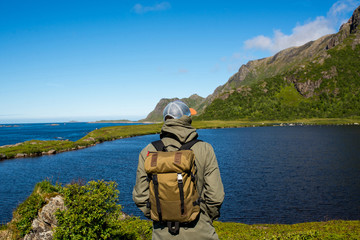 A man with backpack enjoys landscape. Mountain and ocean. Scenic view. Beautiful nature. Green grass, blue sky. Outdoor leisure activity, hiking. Wanderlust. Adventure, lifestyle. Explore North Norway