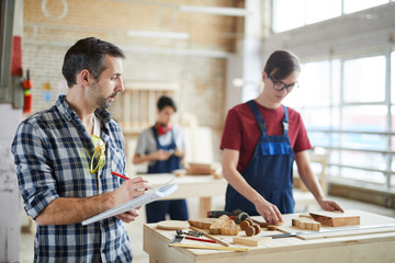 Waist up portrait of mature carpenter holding clipboard d and taking notes while overseeing trainees in workshop class, copy space