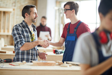 Waist up portrait of mature carpenter shaking hands with young trainee in workshop, copy space
