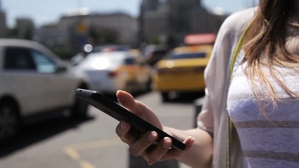 A girl uses a mobile phone on the street on a sunny day against the background of a passing car stream.