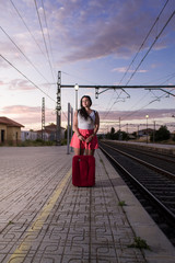 Brunette with pink summer pants waits for the train at the station leaning on her red suitcase.
