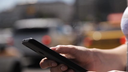 A girl uses a mobile phone on the street on a sunny day against the background of a passing car stream.