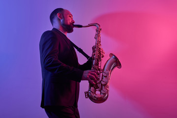 Portrait of professional musician saxophonist man in suit plays jazz music on saxophone, pink...
