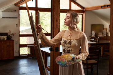 Beautiful talented young woman artist paints a picture with brush and oil on an easel with palette...