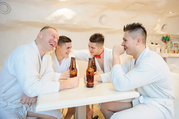 close up photo of 4 men in white gowns sitting around a table and drinking beer after sauna procedures