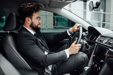 Handsome young bearded driver in full suit with fastening seat belt driving a car.