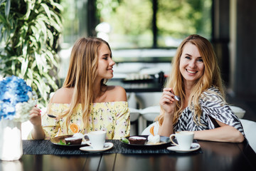 Portrait of mother and blonde daughter talk and smile while drink latte and eating dessert. Spend time happy. Morning breakfast sweets.