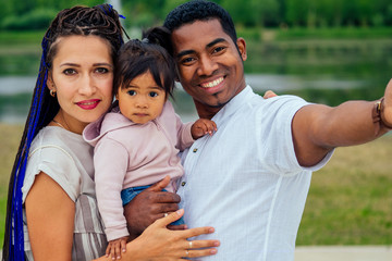 Happy young mixed race transnational interracial couple spending time with their daughter using smartphone making selfie photo camera portrait