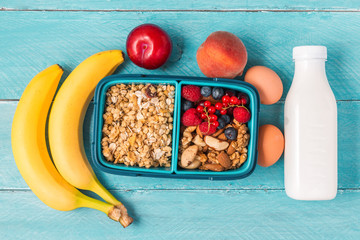 school lunch box with granola, nuts, berries, banana, eggs, peaches and yogurt to make breakfast on blue background