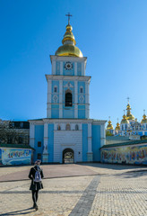 A girl in a skirt standing in front of St Michael's Golden-Domed Cathedral in Kiev. The walls of the cathedral are painted blue and nicely decorated on each facade. Golden domes reflect the sun