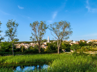 Naklejka premium A little town hiding behind trees and bushes. Lush green color of the plants. Small pond in front. The town in the back is relatively small. Gravel road leading to the center.