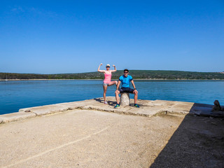 A young couple playing on the shore of Mediterranean Sea, man is sitting on a bollard, and girl is showing off her muscles. Calm sea water. Clean and sunny day. Having fun while active traveling.